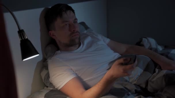 Pria muda Kaukasia yang menderita depresi dan insomnia menjadi marah karena mencoba menghibur dirinya sendiri ketika menonton tv melalui layanan streaming, beralih saluran dengan remote control di tempat tidur di malam hari — Stok Video