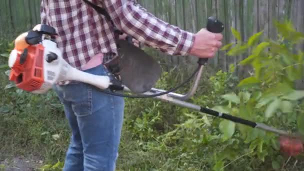 Een grasmaaier die gras maait. Man met oorbeschermers die gras maaien met een benzineheggenschaar. Tuinman maait gras maakt gebruik van gas zeis langs de weg in het dorp. Proces van het snoeien van gazons — Stockvideo