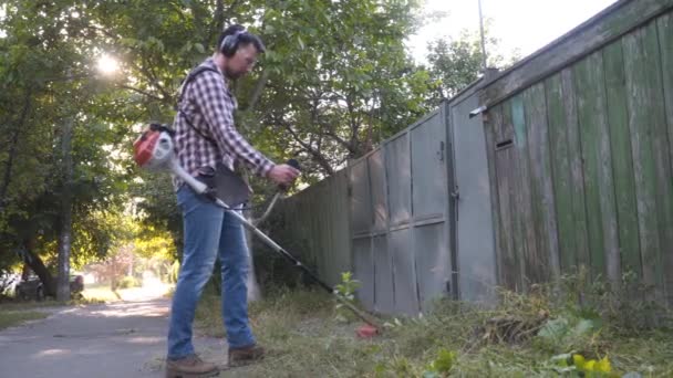 Cortacésped trabajador de corte de césped hombre. Hombre con protectores de orejas segando hierba con recortador de seto de gasolina. Jardinero corta hierba utiliza guadaña de gas a lo largo de la carretera en la aldea. Proceso de recorte del césped — Vídeo de stock