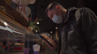Maskeli adam süpermarkette raftaki plastik kutuda suşi seçiyor. Japon yemeği siparişi. Maske yok, giriş yok. Tek kullanımlık plastik konteynırda sağlıklı yiyecekler. Suşi ile beslenme çantası.