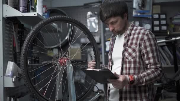Stylowy mechanik rowerowy robi notatki w schowku w warsztacie. Motyw małych firm sprzedających rowery, warsztat rowerowy. Prowadzenie interesów z dokładnością. Naprawiający rowery robi notatki, listy kontrolne — Wideo stockowe