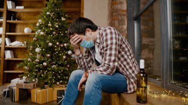Covid 19 yüz maskesi takan yalnız bir adam çok üzgün ve sarhoş, pencerenin ve Noel ağacının yanında oturuyor ve şişeden alkol içiyor. Konik alkol bağımlılığı, karantina sırasında depresyon koronavirüs.