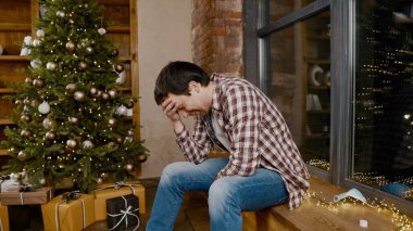 Covid 19 yüz maskesi takan yalnız bir adam çok üzgün ve sarhoş, pencerenin ve Noel ağacının yanında oturuyor ve şişeden alkol içiyor. Konik alkol bağımlılığı, karantina sırasında depresyon koronavirüs