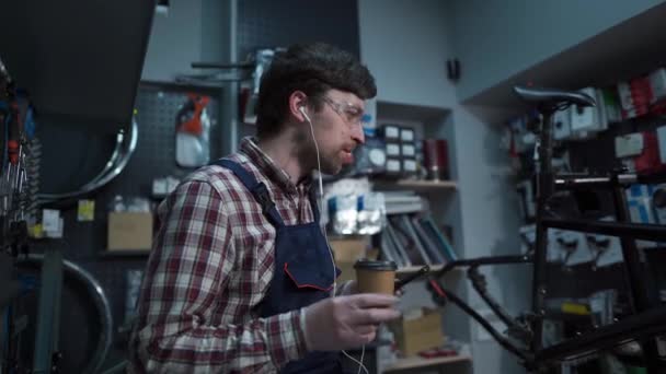 Young caucasian bicycle mechanic listening to music with headphones, drinking coffee and dancing in workshop. Male worker in service uniform for repair bike on coffee break listens music and dances — Stock Video