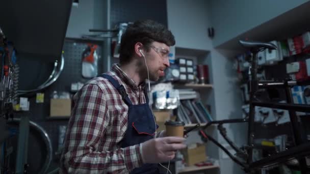 Joven mecánico de bicicletas caucásico escuchando música con auriculares, bebiendo café y bailando en el taller. Hombre trabajador en uniforme de servicio para bicicleta de reparación en coffee break escucha música y bailes — Vídeos de Stock