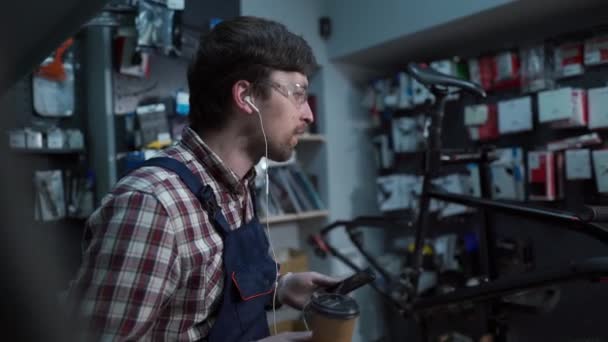 Hombre feliz mecánico de bicicleta en uniforme con sonrisa escucha música con auriculares en el lugar de trabajo, tiempo de descanso café. Trabajador reparador divirtiéndose escuchando música disfrutando de la canción excitada en el concepto de estilo de vida — Vídeos de Stock