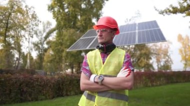 Koruyucu giysiler içindeki erkek işçi, elleri göğsünde yeni kurulmuş bir güneş panelinin arka planına karşı gururla duruyor. Kavramsal yenilenebilir enerji, teknoloji, elektrik, hizmet, yeşil güç
