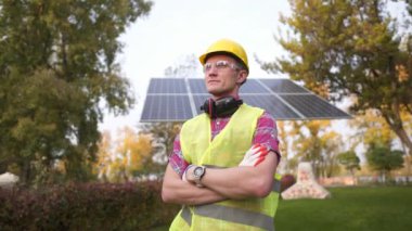 Alternatif enerji kavramı. Koruyucu üniformalı, sarı şapkalı, güneş panelinin yanında poz veren genç ve gururlu bir işçi. Sürdürülebilir temiz enerji. Güneş enerjisi istasyonunda çalışan mutlu tamircinin portresi.