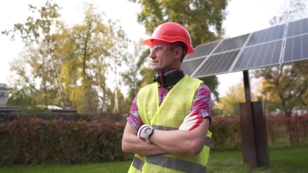 Orgulhoso jovem engenheiro sorri satisfeito com seu trabalho bem sucedido com seus braços cruzados perto da usina solar. Engenheiro fotovoltaico. Conceito de manutenção e instalação de estações solares — Vídeo de Stock