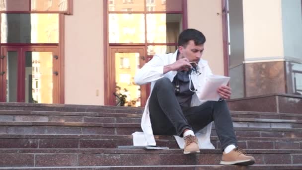 Kardiolog držet a zkontrolovat elektrokardiograf EKG nebo EKG diagram sedí mimo nemocnici na schodech. Mladý doktor student studující pracovní srdce před vyučováním. Vyšetření kardiovaskulárního systému — Stock video