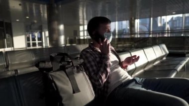 Yorgun, maskeli yolcu boş bir havaalanı terminalinde oturuyor, uçuş ertelenirken telefonla destek alarak konuşuyor. Karantina koronavirüsü sırasında seyahat edin. Uçuş iptal edildi, uçuş reddedildi.