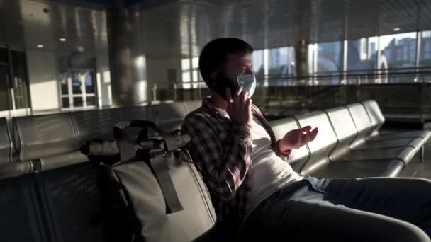 Vermoeide, gemaskerde passagier zit in een lege luchthaventerminal, telefonerend met ondersteuning als de vlucht vertraagd is. Reizen tijdens quarantaine coronavirus. Vluchtannulering, instapweigering in geval van covid 19 — Stockvideo