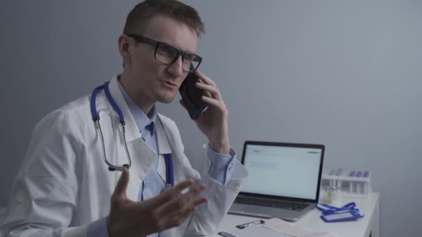 Mosolygó doktor telefonál az asztalnál. Orvos beszél a betegével az irodai klinikán, aki laptoppal ül az asztalnál. Német orvos fehér köpenyben dolgozik kórház, konzultál online telefonon — Stock videók