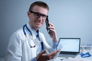 Gülümseyen doktor masasında telefonla konuşuyor. Ofis kliniğindeki hastasıyla konuşan hekim laptopuyla masada oturuyor. Beyaz önlüklü Alman doktor hastanede çalışıyor, internet üzerinden danışmanlık yapıyor..