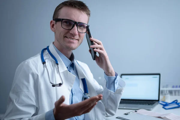 Smiling doctor having phone call at desk. Physicianr talking with his patient in office clinic sitting at table with laptop. German doctor in white lab coat works hospital, consults online by phone.