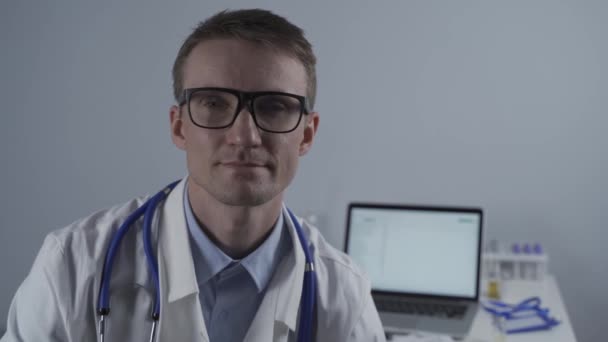 幸せな自信を持った男性専門医の肖像画を閉じます。笑顔の医者はオフィスのカメラを見ると制服を着ている。ハンサムなドイツの笑顔の一般開業医のクローズアップ眼鏡をかけて — ストック動画