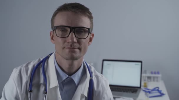 Feliz médico profesional masculino seguro de sí mismo cerca de retrato. Médico sonriente usa uniforme mirando la cámara en el consultorio. Primer plano del guapo médico general alemán sonrisa con gafas — Vídeos de Stock