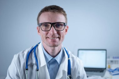 Kendine güveni tam, profesyonel bir doktor portreyi kapatıyor. Gülümseyen doktor ofisteki kameraya bakarken üniforma giyer. Yakışıklı Alman gülüşlü pratisyen hekimin yakın çekimi..
