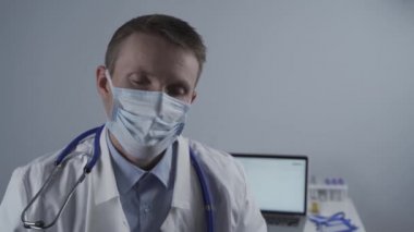 Yorgun genç doktor gözlüklerini ve koruyucu tıbbi maskelerini çıkarıp hastane ofisindeki masaüstü kameralarına bakıyor. Konusu Coronavirus 'taki aşırı yüklenmiş tıbbi sistem, salgın covid 19.