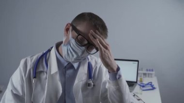 Laboratuvar önlüklü yorgun genç doktor maskesini ve gözlüklerini çıkarıyor, gözlerini ovuşturuyor, yorgunluktan başı ağrıyor ve hastanede ağır vardiyada çalışmaktan dolayı aşırı yorgunluk çekiyor. Doktor ofisteki kameraya yorgun bakıyor.