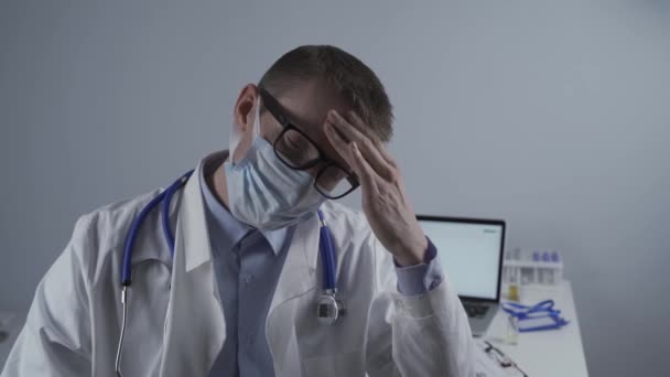 Utmattad ung läkare i labbrock tar av mask och glasögon, gnuggar ögon, har huvudvärk av trötthet och överansträngning från att arbeta i tunga skift på sjukhus. Doktorn ser trött på kameran på kontoret — Stockvideo