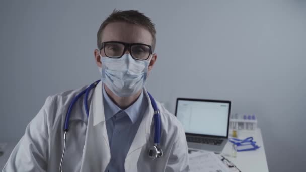 Γερμανός Ευρωπαίος νεαρός γιατρός που κοιτάζει την κάμερα φορώντας γυαλιά και μάσκα στο γραφείο της κλινικής στο χώρο εργασίας με κουρασμένη εμφάνιση. Το θέμα είναι η σκληρή δουλειά των ιατρών κατά τη διάρκεια της πανδημίας coroavirus — Αρχείο Βίντεο