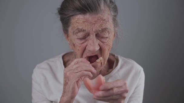 Mycket gammal kaukasisk senior kvinna 90 år med djupa rynkor och grått hår passade tandprotes på grå vägg bakgrund i studio. Äldre kvinnor sätter protes. Falska tänder. Tandvård — Stockvideo