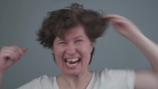 Biała kobieta jest szalenie chwytana za włosy, traci zęby, śmieje się i krzyczy patrząc na kamerę w studio na szarym tle. Wściekła kobieta rozdzierająca włosy. Szalony nastrój — Wideo stockowe