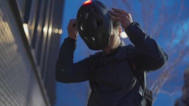 Bisikletli erkek teslimatçı, akşam bisikletli alıcıya sipariş vermeden önce arka sinyal lambasıyla kask takıyor. Şehirdeki işten eve gidip bisiklet kaskı takan bir adam.