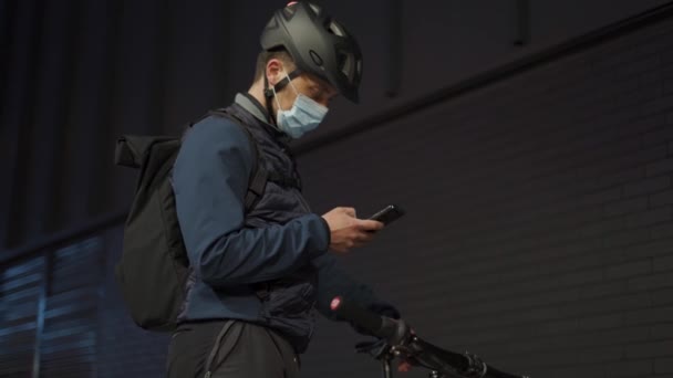 ヘルメットを着用した自転車の配達人と保護医療マスクは、市内の夜に携帯電話の方向をチェックします。コロナウイルスパンデミック時のオンラインストアからの商品の配達。バイク宅配便 — ストック動画