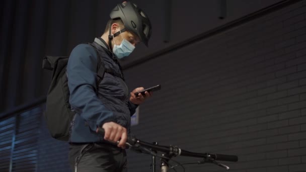 Кавказский курьер в медицинской маске и шлеме стоит на улице на велосипеде и отправляет сообщение по телефону в ночном городе. Велогонщик печатает на мобильном телефоне на улице. Концепция поставки ковид 19 — стоковое видео