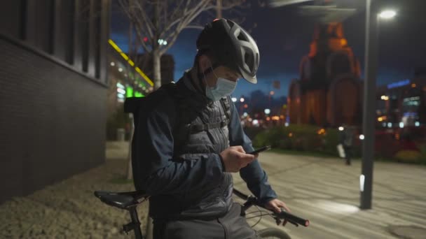 El repartidor en bicicleta con casco y máscara médica de protección comprueba las instrucciones en el teléfono móvil por la noche en la ciudad. Entrega de productos de tiendas en línea durante la pandemia de coronavirus. Correo de bicicleta — Vídeos de Stock