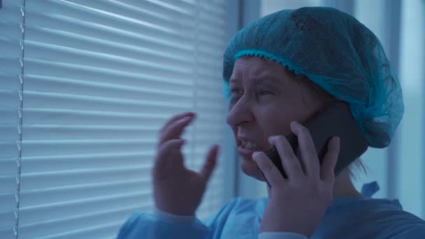 Szomorú női beteg orvosi ruhában, telefonon beszél és sír egyedül a kórházban, női várakozó orvos az ablakban a kórházban, stresszes, sír és mobiltelefon baleset miatt — Stock videók