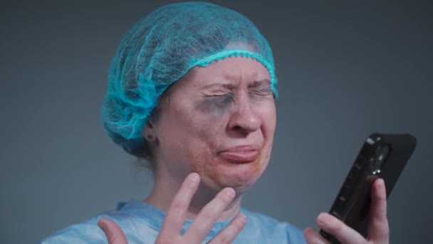 Mujer paciente con bata de protección médica azul desechable llorando y llamando a la compañía de seguros en el hospital de emergencia. Mujer con lesiones faciales después de la violencia en la clínica llama al servicio social — Vídeos de Stock