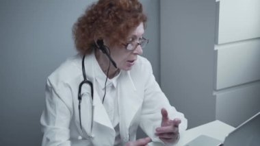 Çevrimiçi tıbbi tedavi uygulayan kadın sağlık çalışanı. Tıbbi asistan bilgisayardan uzak hastayı ararken kulaklık takıyor. Teletıp konsepti. Çevrimiçi doktor randevu uygulaması sanal toplantı