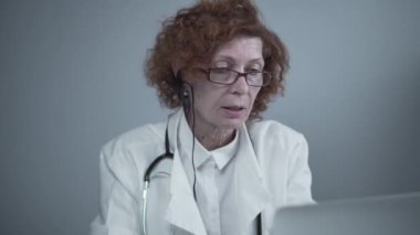 Beyaz önlüklü ve kulaklıklı kadın kıdemli doktor bilgisayarda video konferans için çevrimiçi video çağrı uygulaması kullanıyor. Hastalara uzaktan tıbbi bakım, online danışma