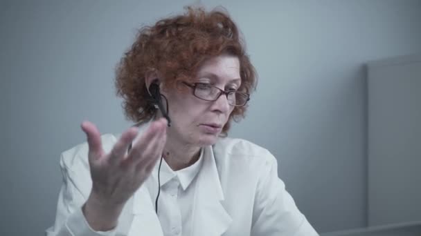 Beyaz önlüklü ve kulaklıklı kadın kıdemli doktor bilgisayarda video konferans için çevrimiçi video çağrı uygulaması kullanıyor. Hastalara uzaktan tıbbi bakım, online danışma — Stok video