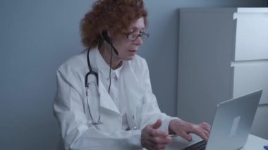 Çevrimiçi tıbbi tedavi uygulayan kadın sağlık çalışanı. Tıbbi asistan bilgisayardan uzak hastayı ararken kulaklık takıyor. Teletıp konsepti. Çevrimiçi doktor randevu uygulaması sanal toplantı