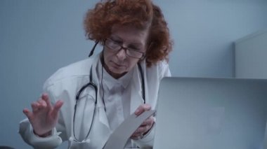 Beyaz önlüklü ve kulaklıklı kadın kıdemli doktor bilgisayarda video konferans için çevrimiçi video çağrı uygulaması kullanıyor. Hastalara uzaktan tıbbi bakım, online danışma