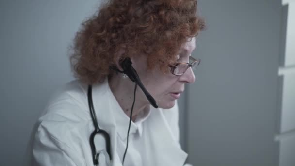 Beyaz önlüklü ve kulaklıklı kadın kıdemli doktor bilgisayarda video konferans için çevrimiçi video çağrı uygulaması kullanıyor. Hastalara uzaktan tıbbi bakım, online danışma — Stok video