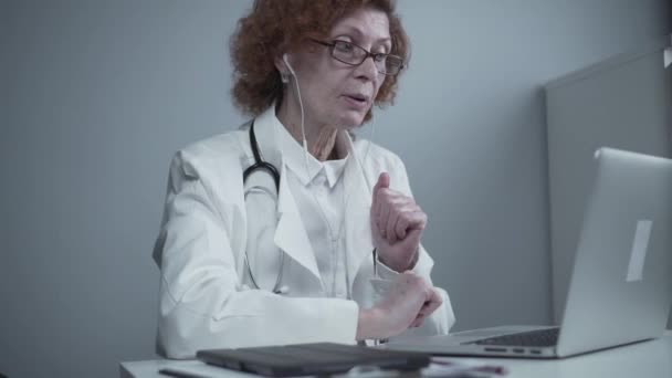 Médecin thérapeute principale en manteau blanc et casque parlant visioconférence sur ordinateur portable au bureau en utilisant l'application d'appel vidéo en ligne. Soins médicaux à distance pour les patients, consultation en ligne — Video