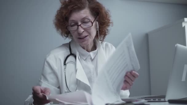 穿着白色外套和耳机的女高级医生治疗师在办公室的笔记本电脑上使用在线视频通话应用程序进行交谈视频会议。为病人提供远程医疗，在线咨询 — 图库视频影像