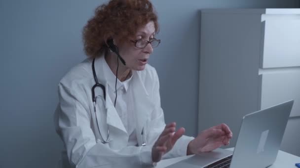 Çevrimiçi tıbbi tedavi uygulayan kadın sağlık çalışanı. Tıbbi asistan bilgisayardan uzak hastayı ararken kulaklık takıyor. Teletıp konsepti. Çevrimiçi doktor randevu uygulaması sanal toplantı — Stok video