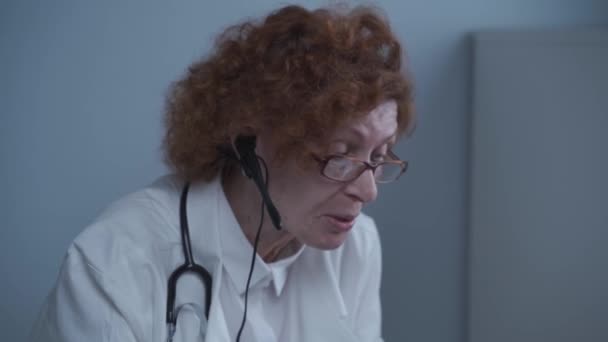 ホワイトコートの女性シニアドクターセラピストとオンラインビデオ通話アプリケーションを使用してオフィスでラップトップでビデオ会議を話してヘッドセット。患者の遠隔医療、オンライン相談 — ストック動画