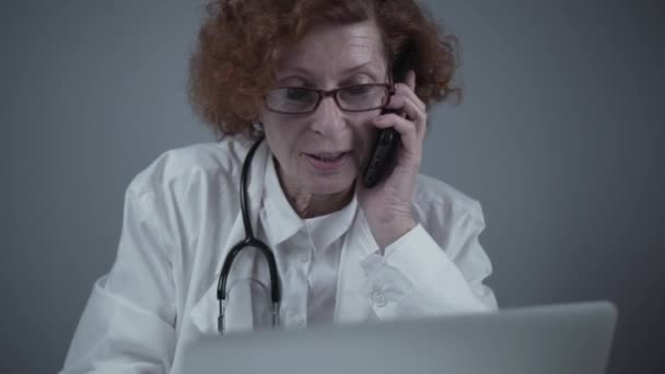快乐而成熟的女医生用笔记本电脑工作，在办公室里通过电话交谈。和病人有关。医药、人和保健概念。全科医生通过智能手机与同事交谈 — 图库视频影像
