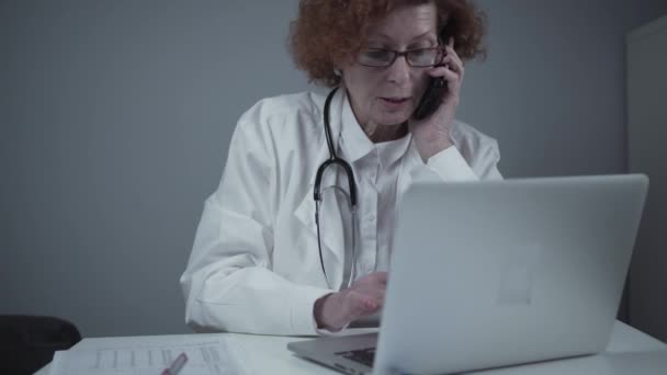 Médico caucásico de mediana edad sentado en su consultorio usando el teléfono y trabajando en el portátil. Médico general de edad avanzada hablando por teléfono con el paciente mientras trabaja en la computadora en la clínica — Vídeo de stock
