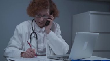 Orta yaşlı, beyaz kadın doktor danışma odasında oturuyor ve bilgisayarla çalışıyor. Klinikte bilgisayarla uğraşırken hastayla telefonda konuşan yaşlı pratisyen.