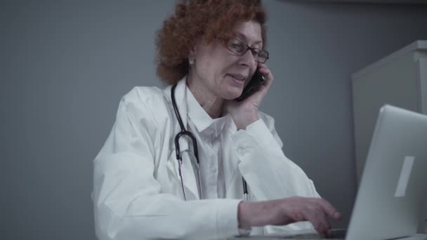 Eine kaukasische Ärztin mittleren Alters sitzt in ihrem Sprechzimmer und arbeitet am Laptop. Älterer Allgemeinmediziner telefoniert mit Patient, während er in der Klinik am Computer arbeitet — Stockvideo