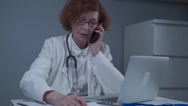 Caucasian senior female doctor physician talking on mobile at work. Healthcare professional answering call giving remote consultation on smart phone make appointment notes on laptop in office — Stock Video