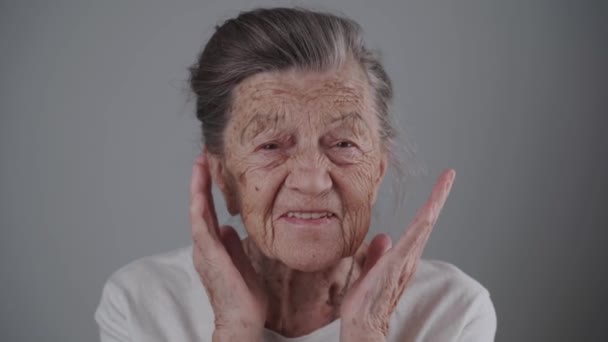 Közelkép elégedett kaukázusi idős nő nézi a kamerát mosolyogva és nevetve szürke háttér. Egy 90 éves nagymama portréja, aki boldog a stúdióban. Műfogsor idős személyek számára — Stock videók